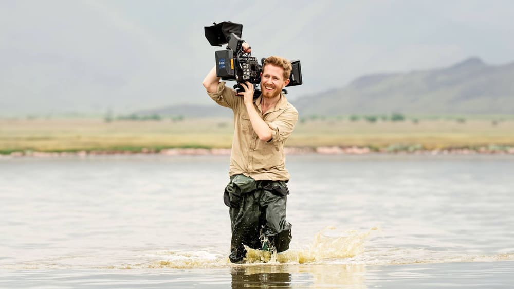 Sebastian wading through shallow water with a camera on his shoulder