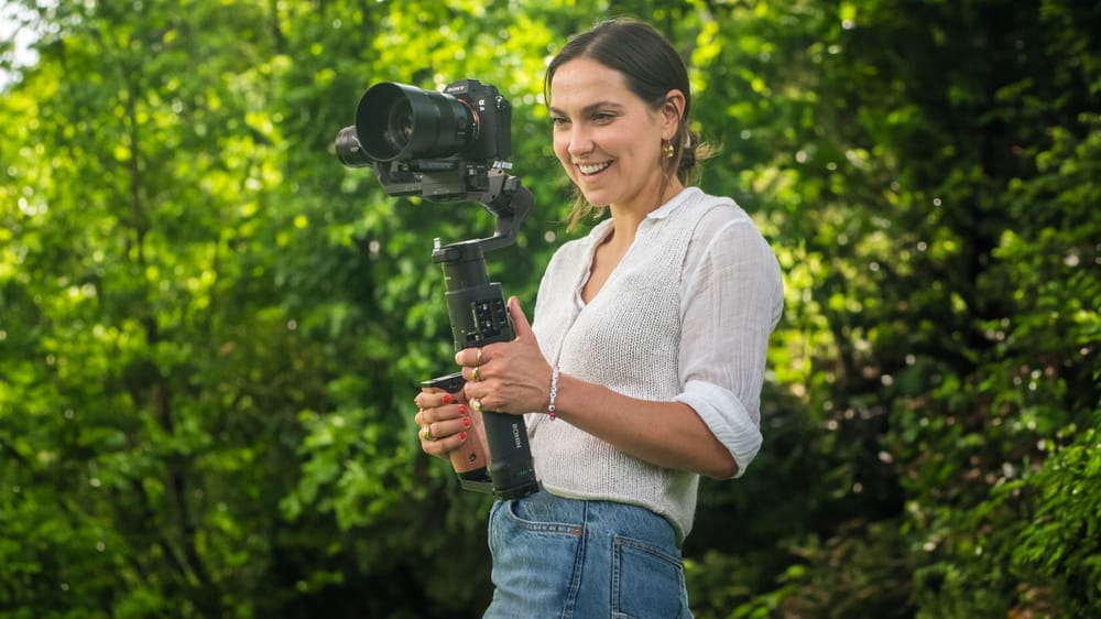 A woman smiling as she films with a camera on a handheld gimbal in woodland