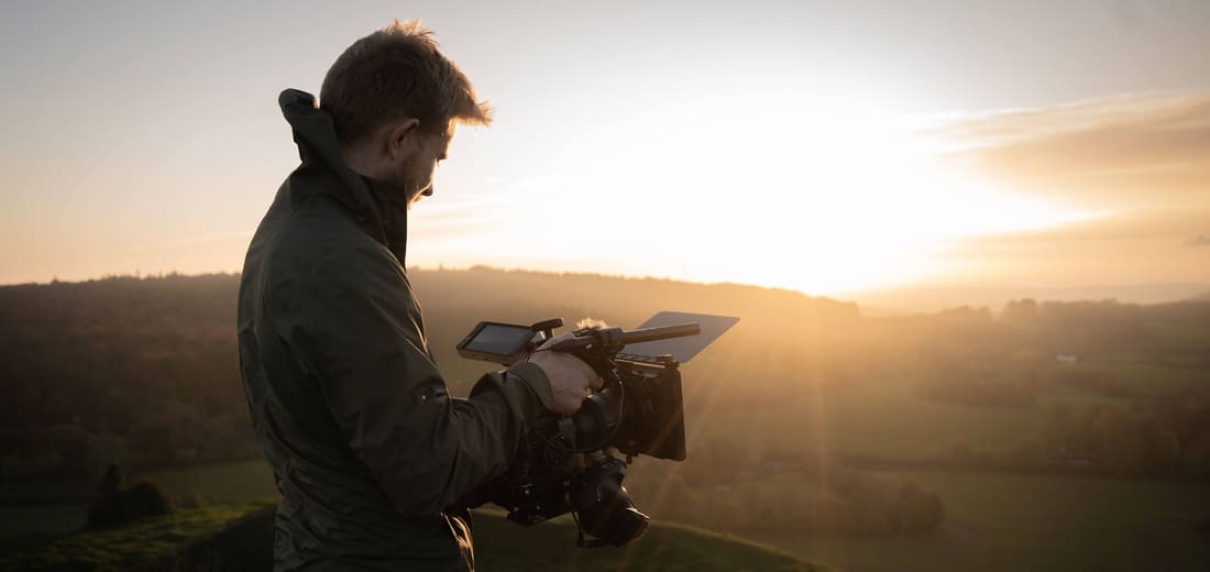 A filmmaker with a camera looking out over hills at sunset