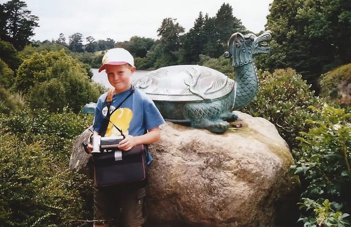 Sebastian aged nine, holding a camcorder, standing beside a bronze turtle statue