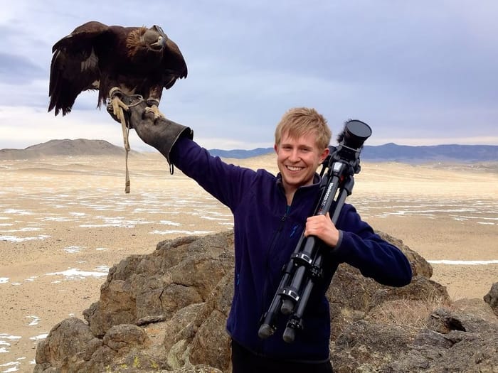 Sebastian in Mongolia, holding a golden eagle on one arm and a tripod over his shoulder