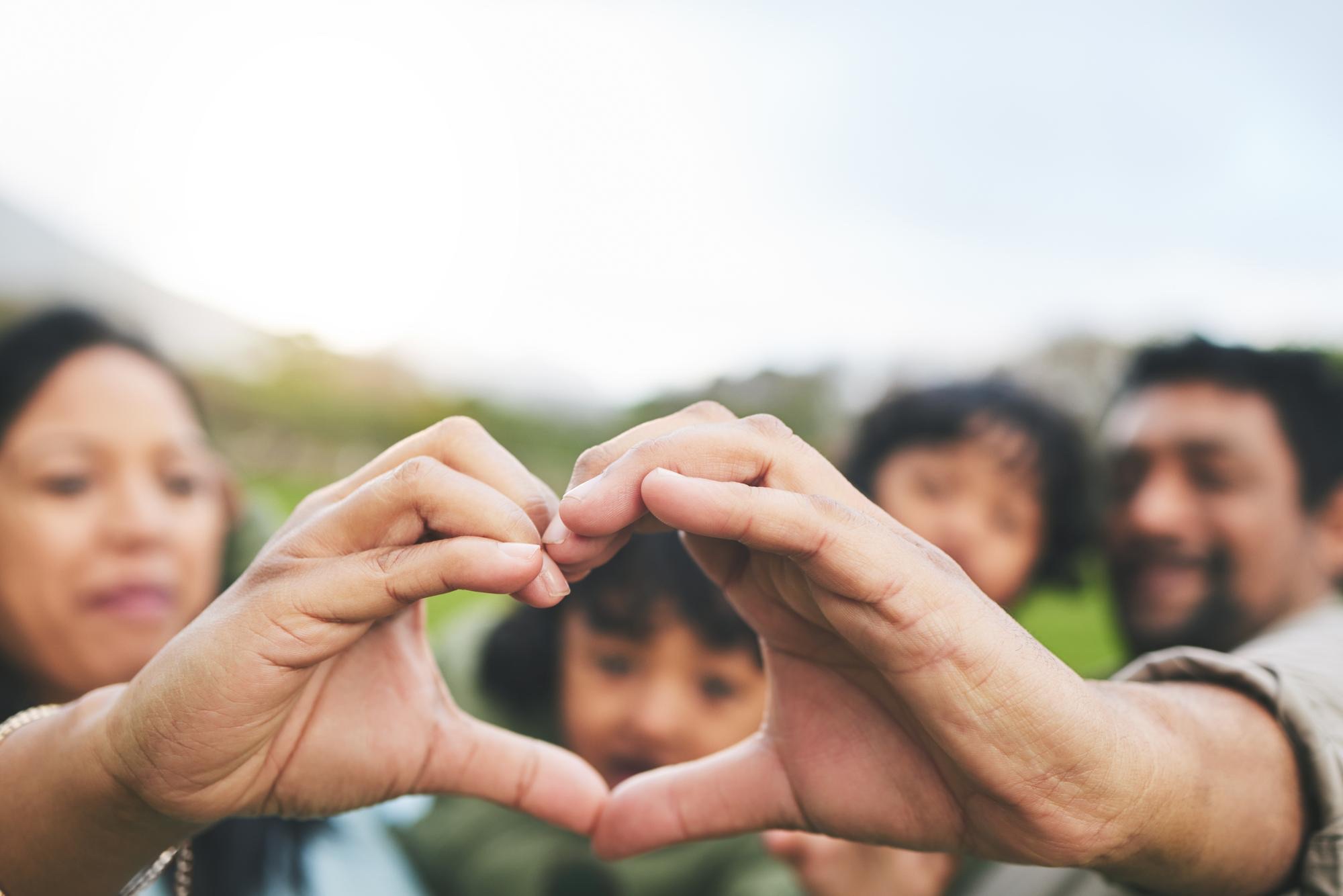 A family forms a heart shape with their hands outdoors, two children and their parents softly out of focus behind