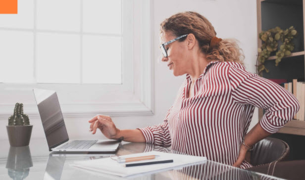 A woman sits at her desk with her hand to her lower back, indicating back pain.