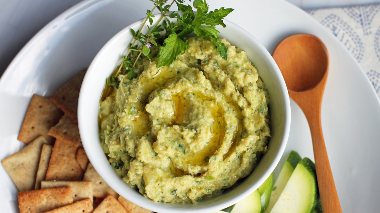 pale green artichoke dip with a sprig of thyme and mint, surrounded by almond flour crackers and fresh zucchini slices on a white plate, with a wooden serving spoon