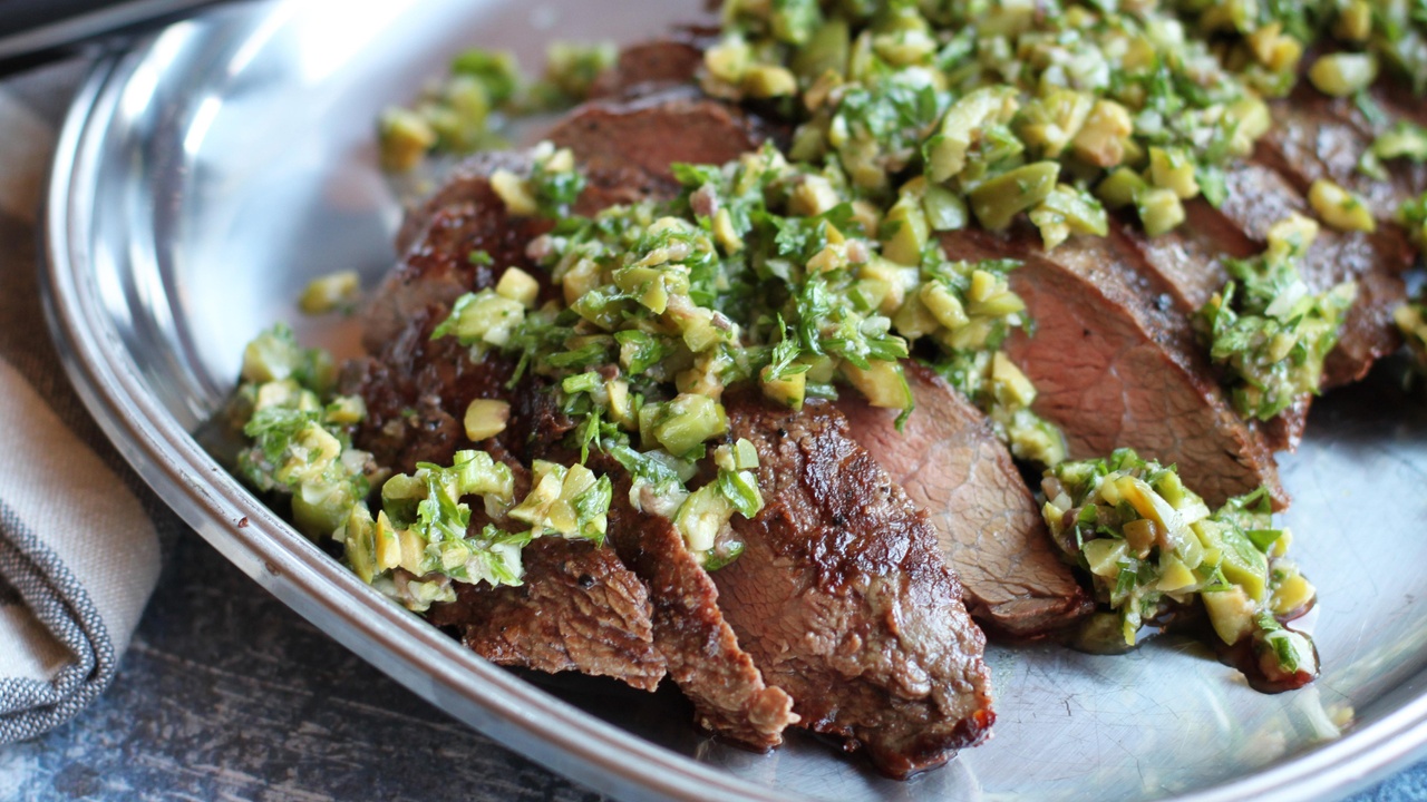 sliced flat iron steak topped with a rustic parsley garlic and olive relish on a silver platter