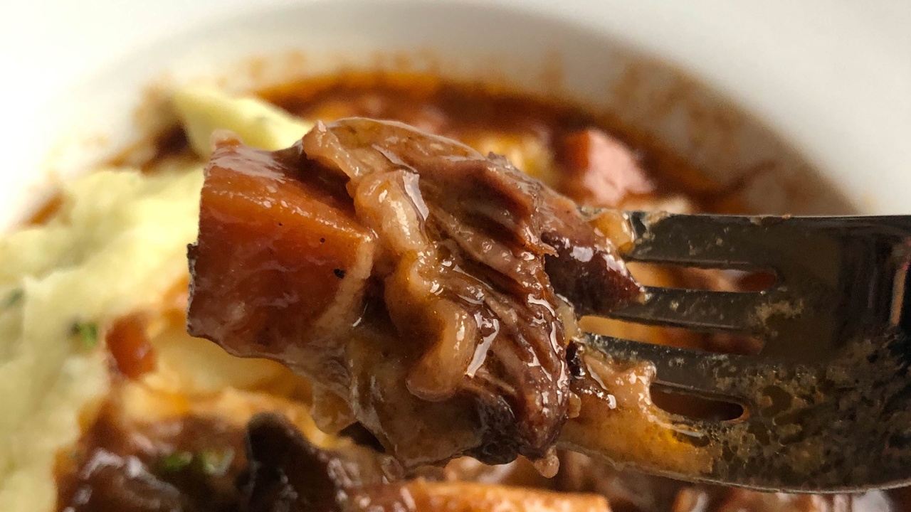 classic braised short ribs atop a cauliflower and white sweet potato mash with cubes of carrot, a rich brown gravy, and fresh parsley on top. They're serveed in a shallow white bowl with a wide rim. 