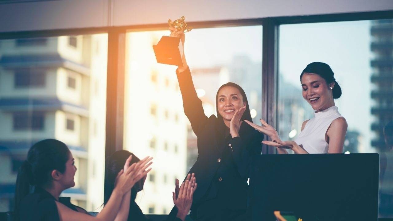 Women in business suit and setting holding up a trophy in front of her co-workers.