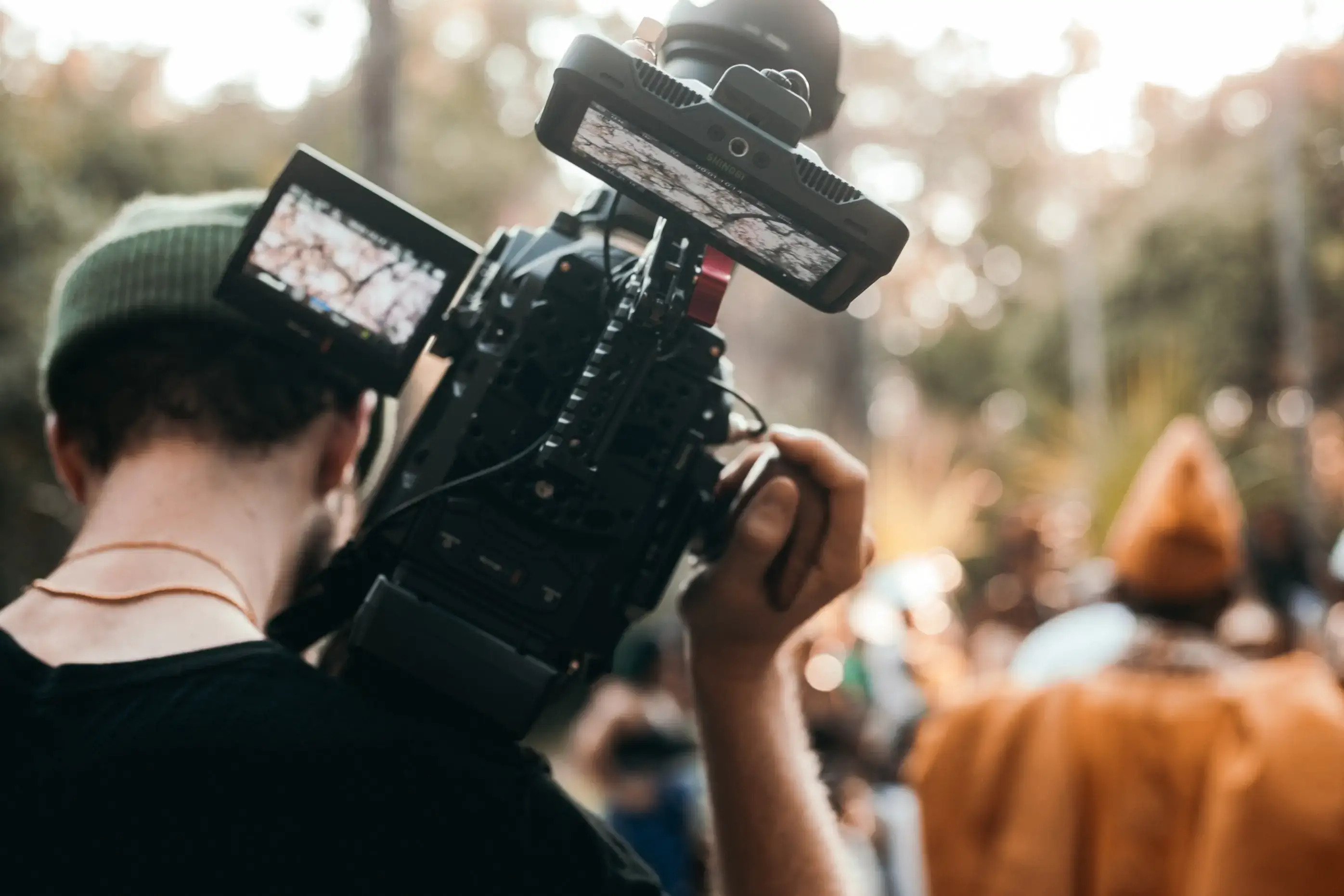 Person wearing a beanie carrying a film camera on his shoulder. The person is facing away from the camera