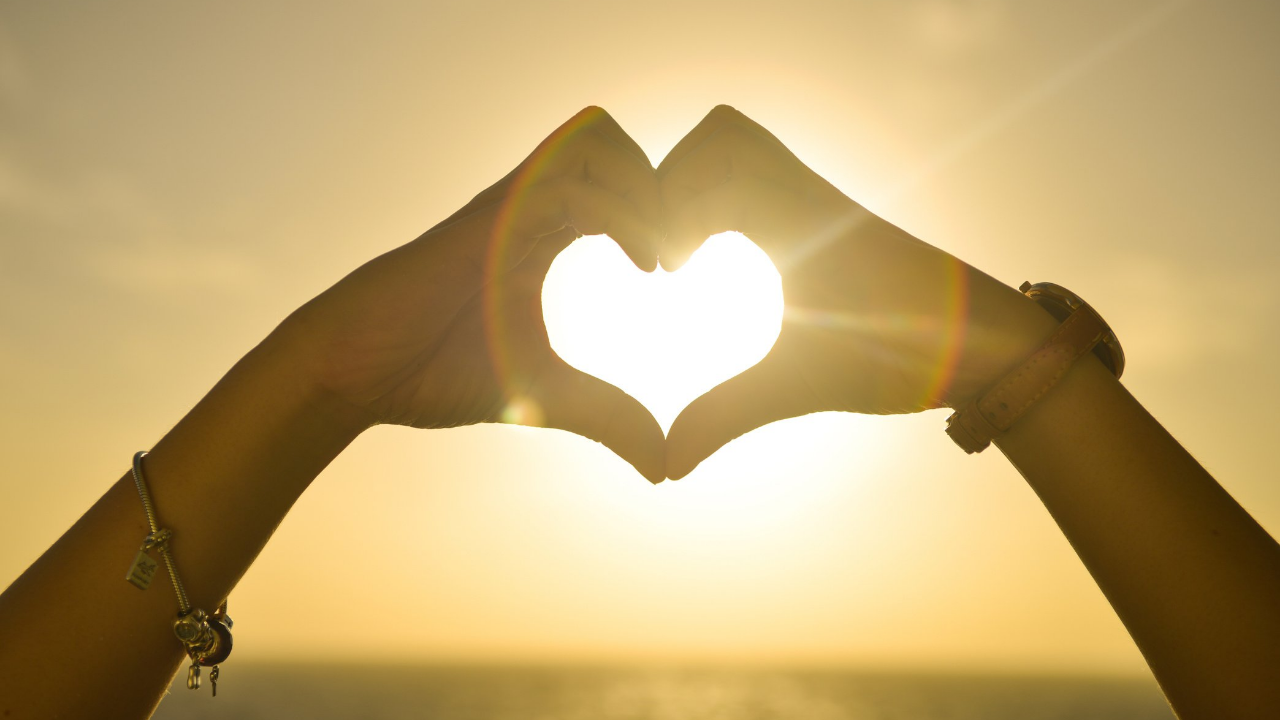 Two hands making a heart shape in front of a sunset.