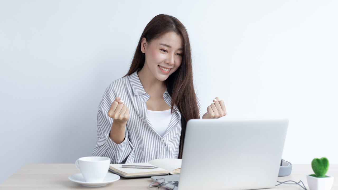 Asian Woman sitting at a table with a laptop, smiling and  saying yes with her hands in front of her.