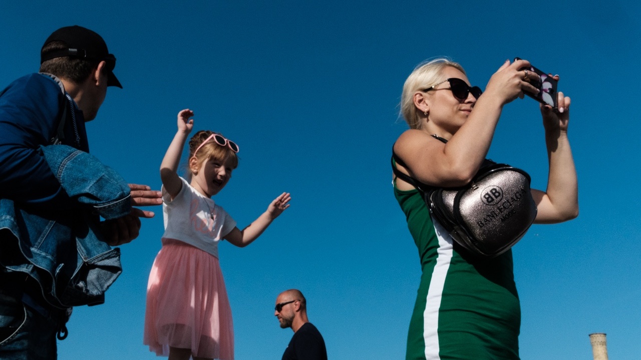 street photography of a little girl in a pink skirt standing on a wall with her arms up in the air behind a woman in a green dress taking a photograph