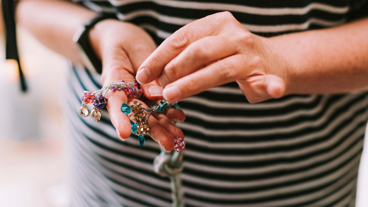 Two hands holding and stitching some silver, turquoise, fuchsia and copper beadwork with a stripy navy and white dress behind