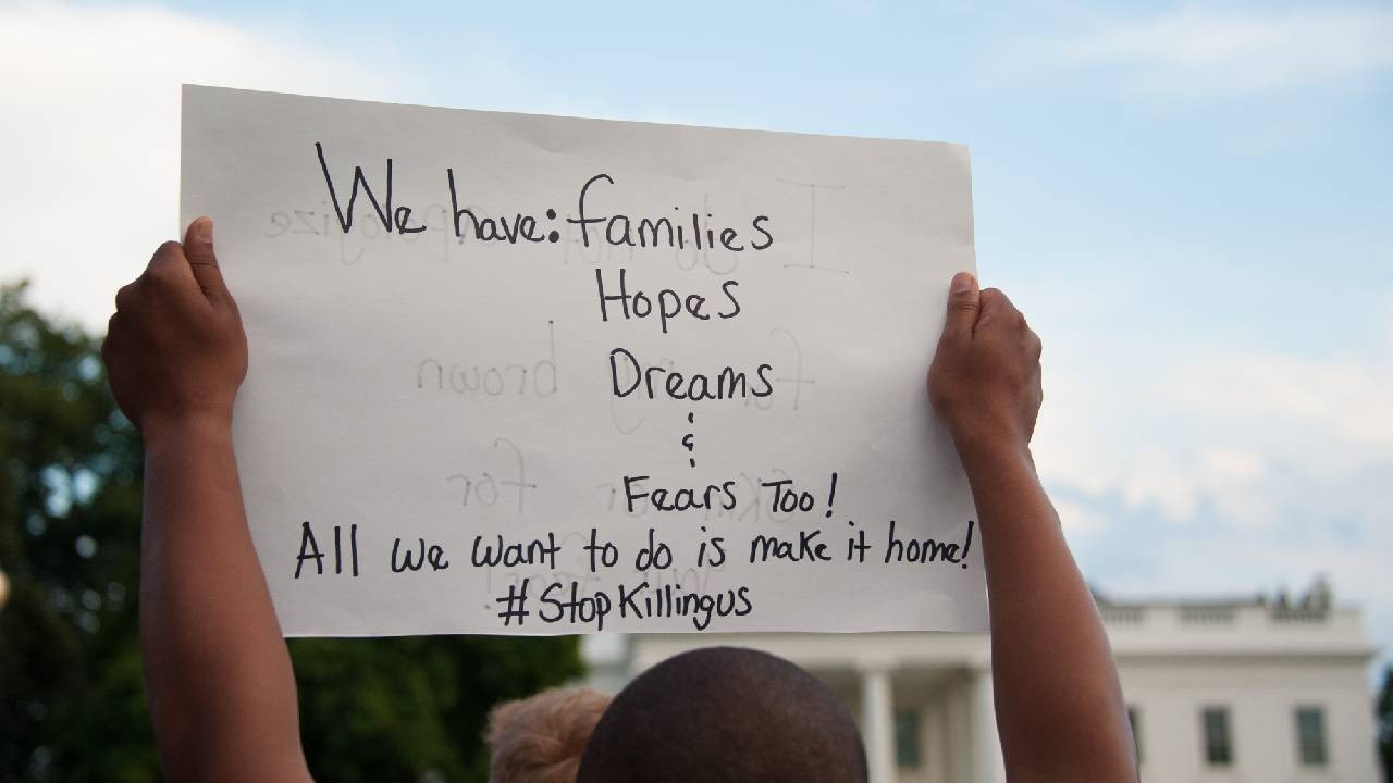 man holding a sign that reads We have families, hopes, dreams, and fear, too! All we want to do is make it home! #StopKillingUs