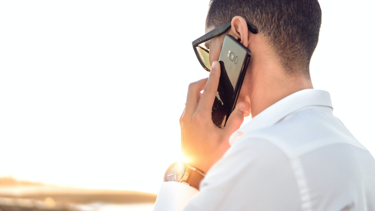 Viewed from behind, a man speaks on a cell phone out doors in the bright sun. He is wearing sunglasses and a watch on his left hand.