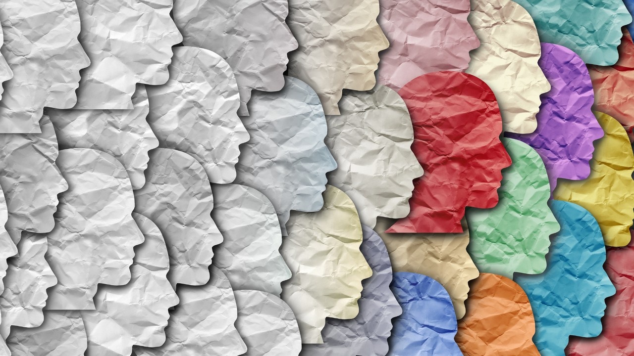 The image depicts a collection of silhouettes of human heads in profile view, arranged in rows and columns. Each silhouette appears to be made out of crumpled paper, and they vary in color across a spectrum from grayscale to vibrant hues. This visual might be interpreted as a representation of diversity and individuality in humanity, with the crumpled paper texture adding an element of complexity or depth to each profile, perhaps symbolizing the multifaceted nature of human personality and identity. The arrangement in a grid could suggest the idea of society or a community where each individual, despite their unique traits, forms part of a larger collective.