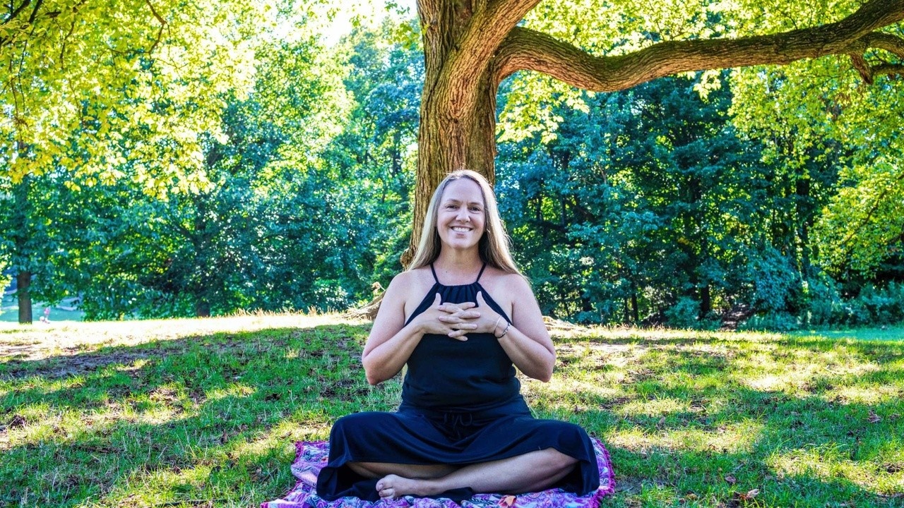 Kate Lynch sitting under a tree on a purple shawl, smiling and demonstrating vajrapradama mudra