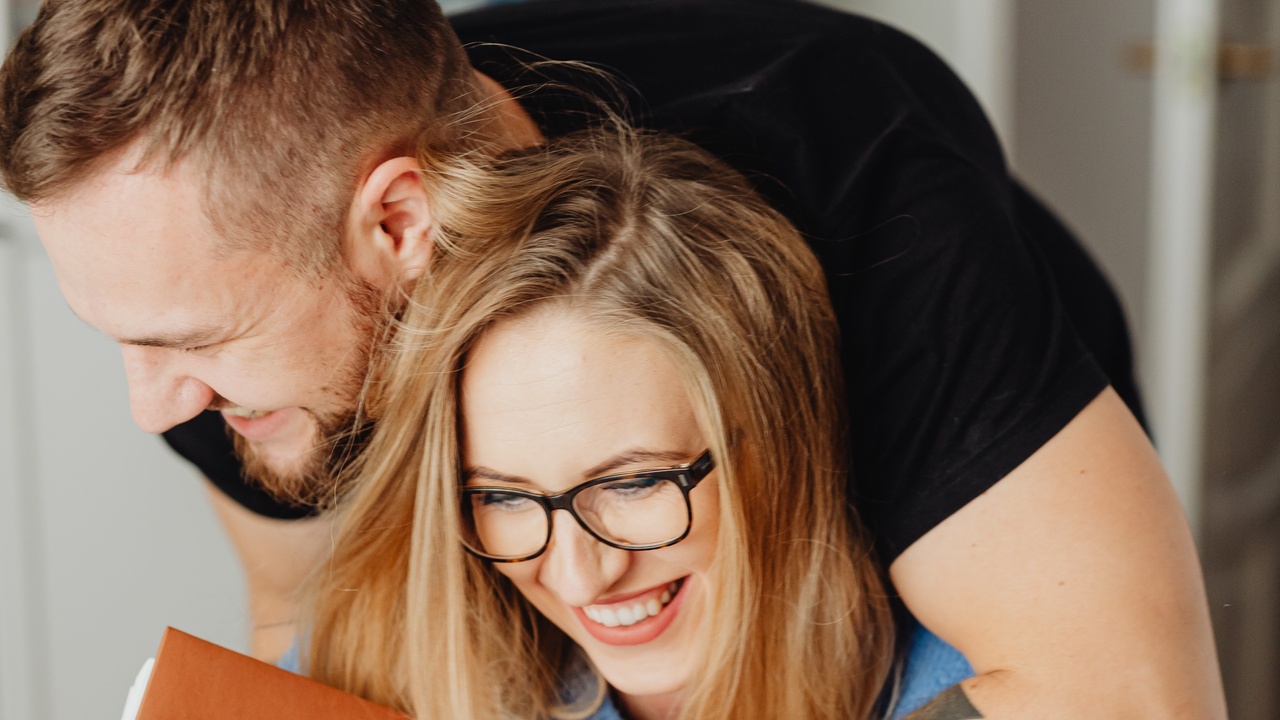 Light skinned couple smiling while masc presenting person hugs a femme presenting person from behind.