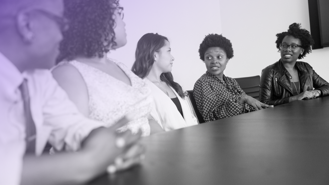 Five people sitting together on one side of the table. They are all looking at one person as they speak, with their hand touching the table.