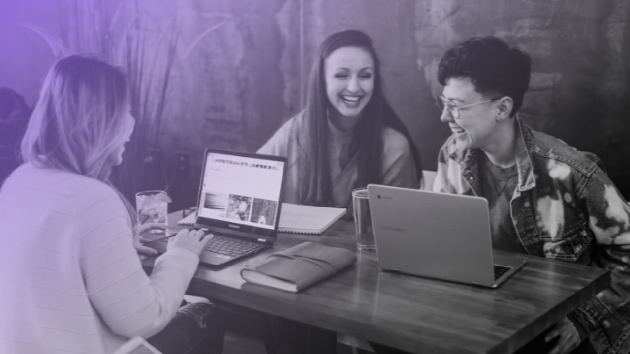 Three people sitting at a large wooden table with laptop open. They are smiling and laughing together.