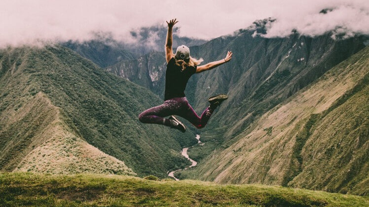 A lady with her back to you jumping in the air, arms up while stood on a mountain