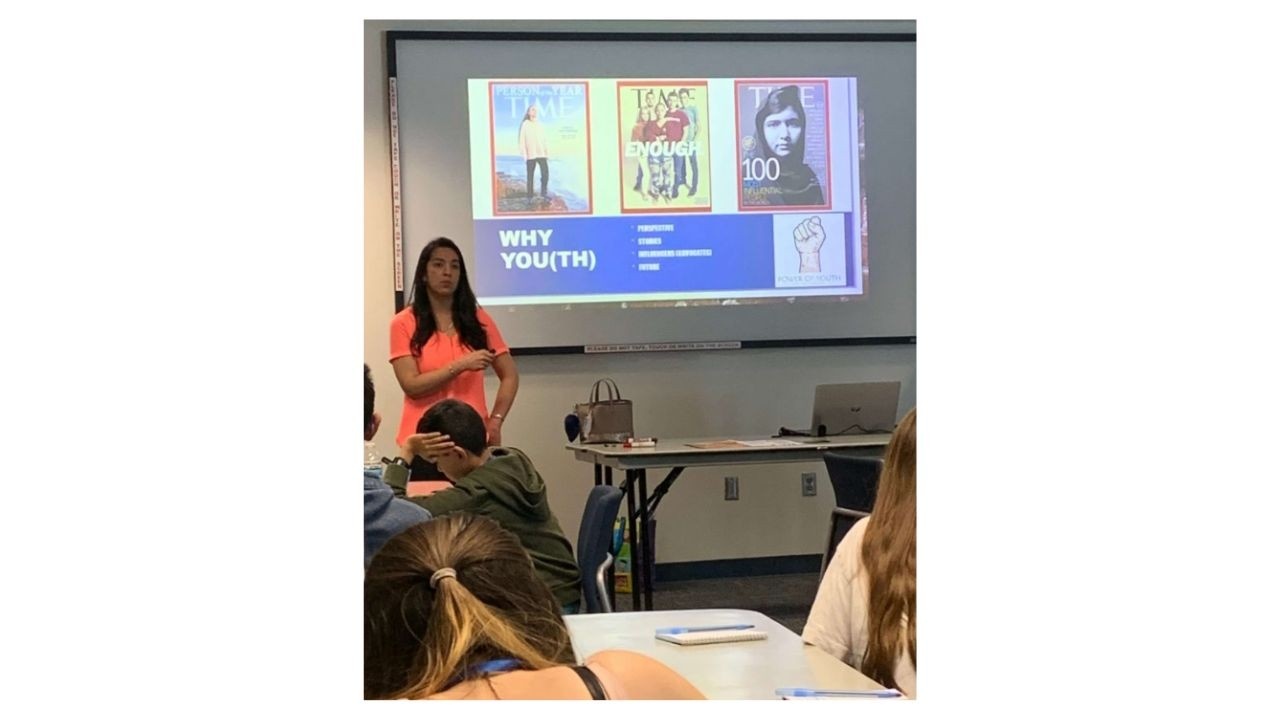 An adult in a bright orange dress is presenting to a classroom of youth and standing in front of a projector screen with a picture of various magazine ads. The slide text says, 