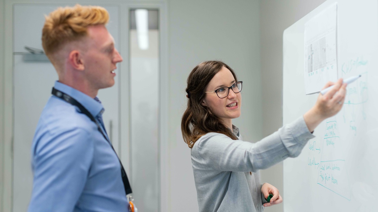 A man watching a woman write on a whiteboard organizing her presentation