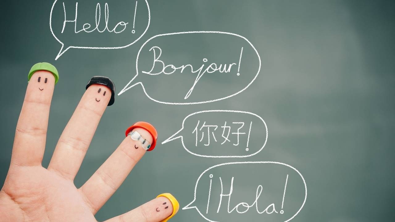 4 fingers with faces drawn on and wearing little hats. Speech bublbles drawn on a blackboard in the background say hello in different languages,