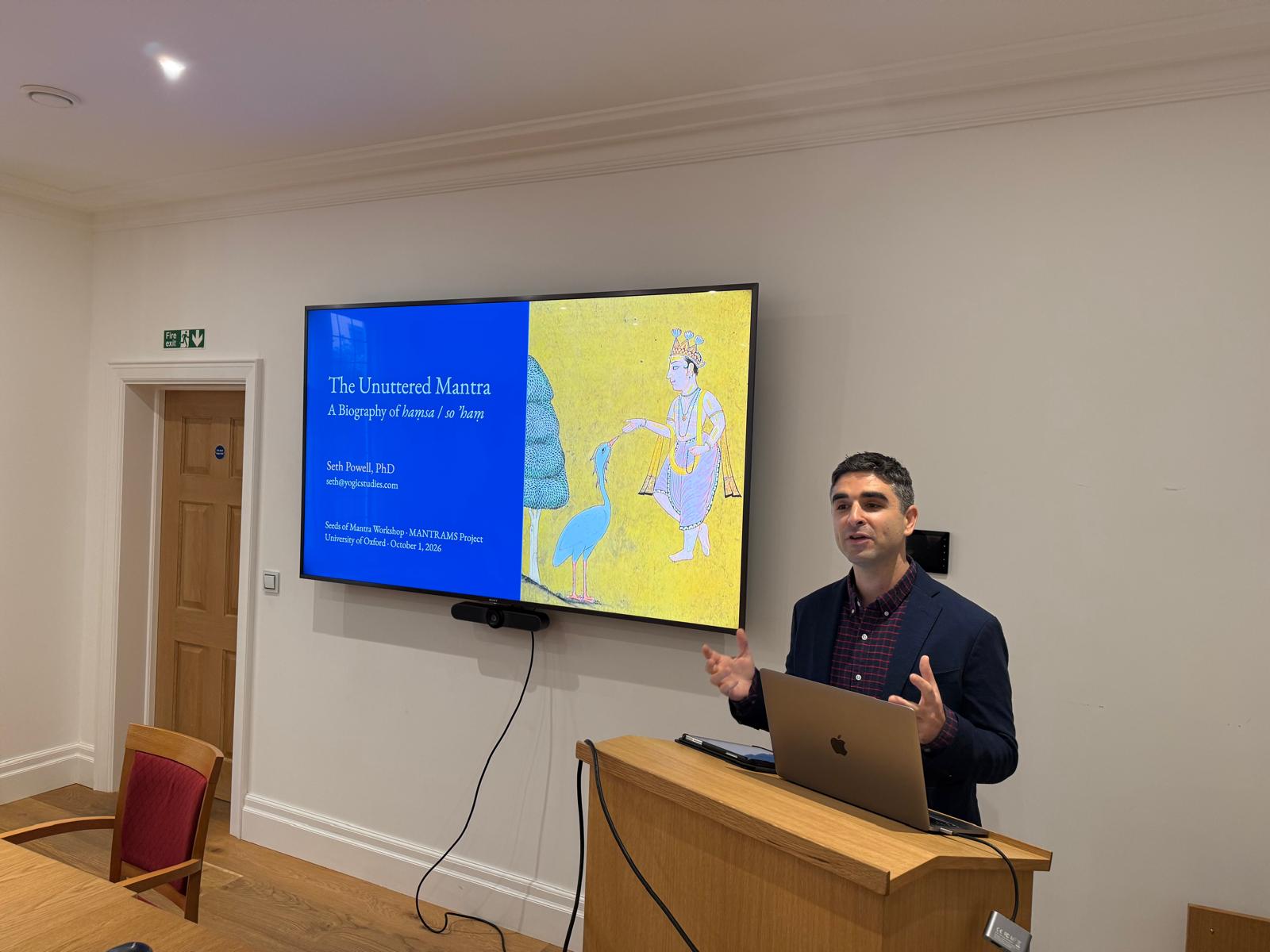 Dr. Seth Powell presenting at a lectern beside a slide reading The Unuttered Mantra: A Biography of haṃsa / so ’haṃ, Seeds of Mantra Workshop, University of Oxford, with a painting of a crowned figure feeding a blue bird
