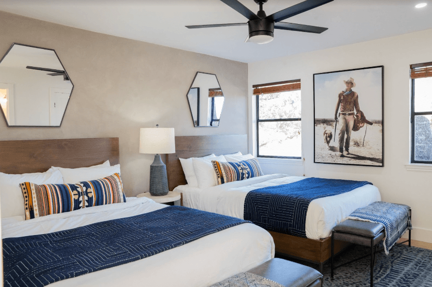 Two beds with white comforters and blue blankets with patterned pillows and two mirros on the wall with John Wayne image hung.