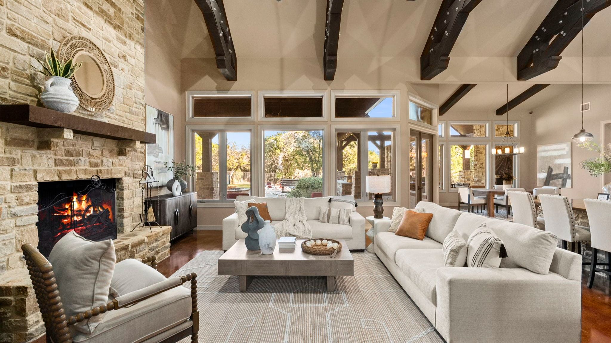 Bright, staged living room with vaulted ceilings and exposed dark wood beams, a stone fireplace with a lit fire, large windows, and neutral-toned sofas arranged around a central coffee table.