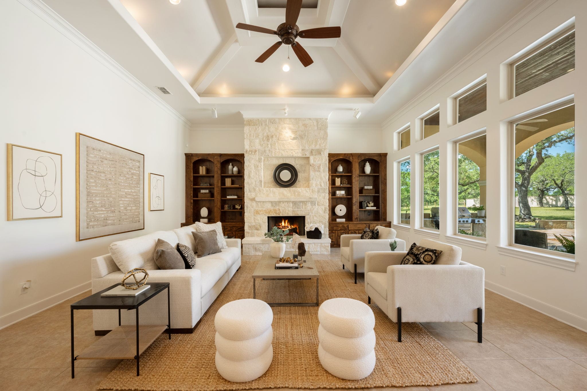 A bright and spacious living room features a high, white tray ceiling with a dark wood ceiling fan at the center. The focal point of the room is a textured stone fireplace with a lit fire, flanked by built-in dark wood bookshelves. Two white sofas and two matching white armchairs are arranged around a gray rectangular coffee table on a natural fiber area rug, complemented by two white textured ottomans in the foreground. A wall of large windows lets in abundant natural light and looks out onto green trees.