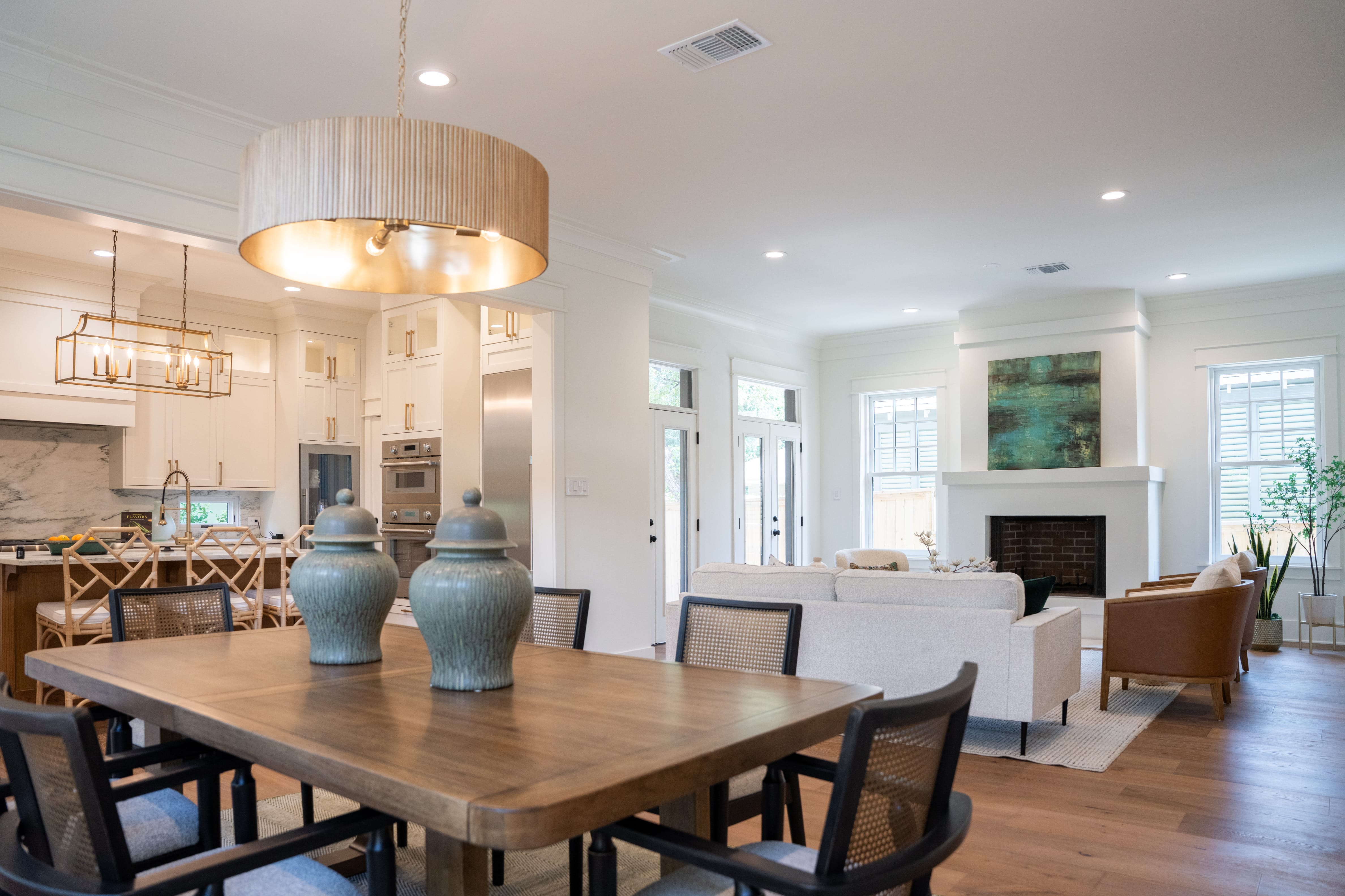 A wide view of a modern, open-concept home transitioning from the dining area to the living room and kitchen. In the foreground, a large wooden dining table is surrounded by black chairs with woven cane backs, highlighted by two large, decorative grey ceramic ginger jars in the center. An oversized, drum-shaped fluted wood pendant light hangs above. In the background, the living room features a cream sofa, leather armchairs, and a white fireplace topped with a teal abstract painting. To the left, a bright white kitchen with built-in appliances and a marble backsplash is visible under a geometric brass chandelier.