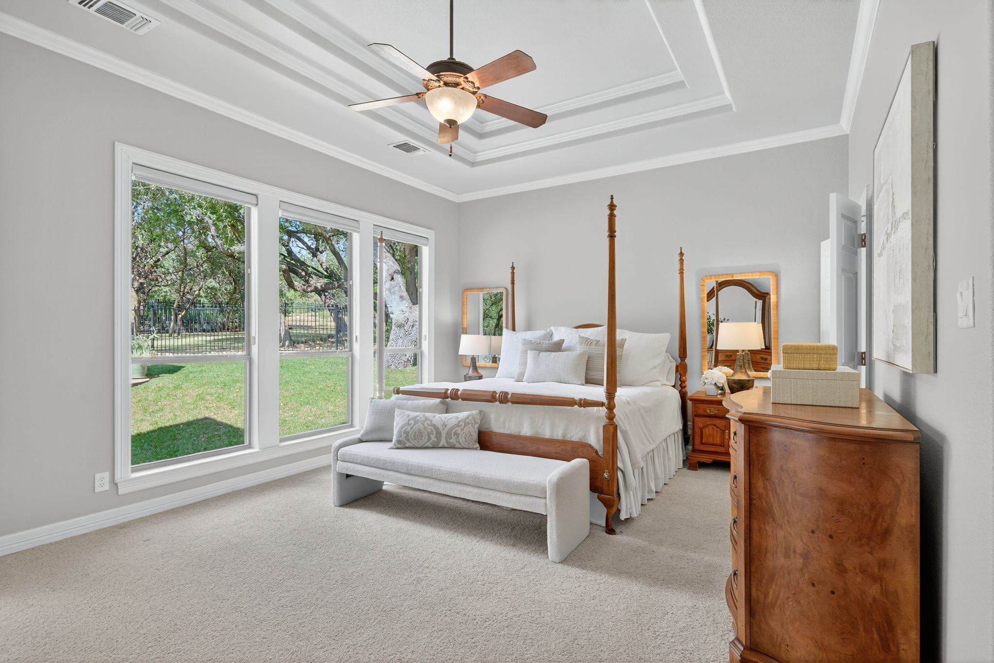 Bright, spacious bedroom with a wooden four-poster bed, upholstered bench, and neutral decor. Large windows line one wall, letting in natural light and showing a green backyard, with a tray ceiling and ceiling fan overhead.