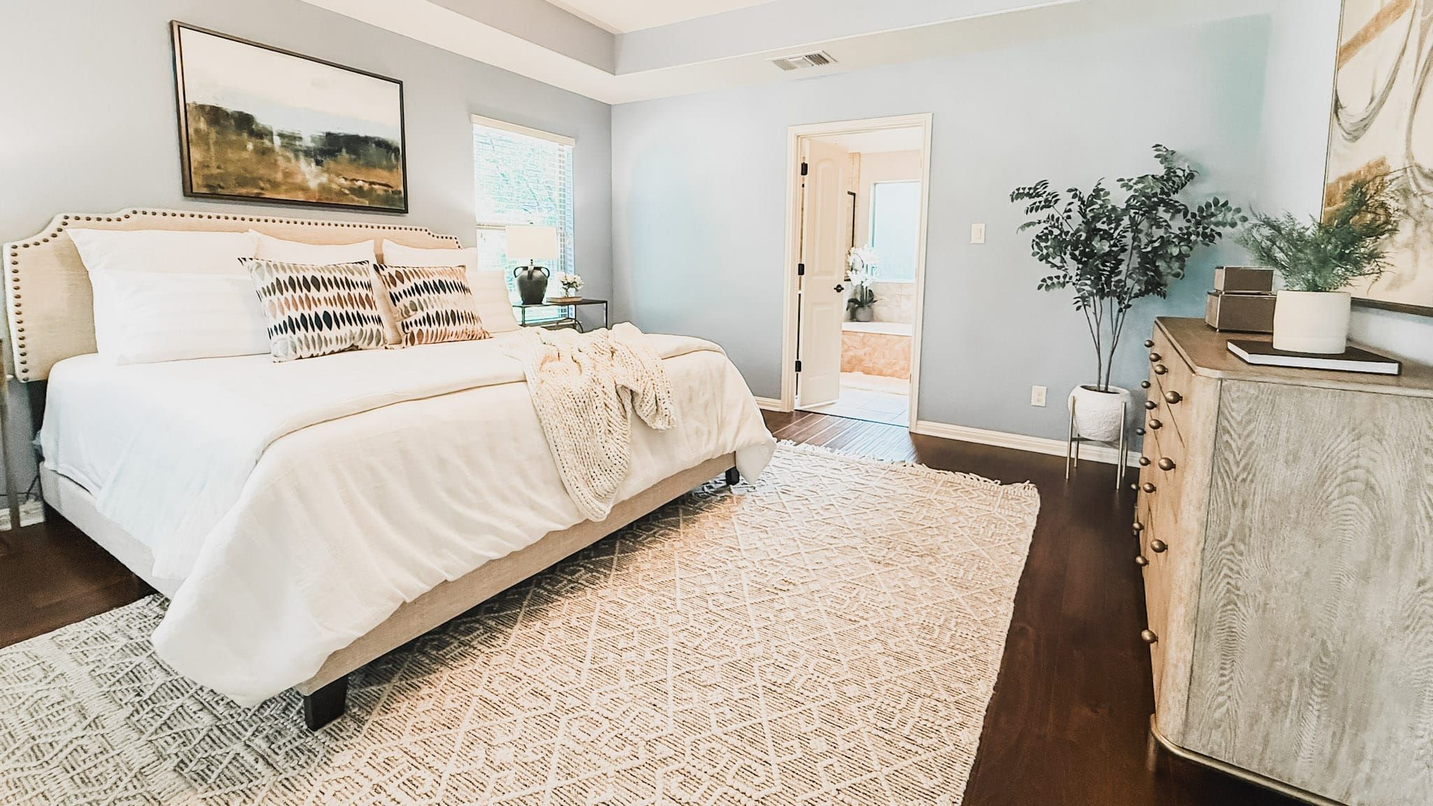 Bright, professionally staged bedroom with a neatly made bed, neutral bedding, hardwood floors, an area rug, wall art, and a potted plant.