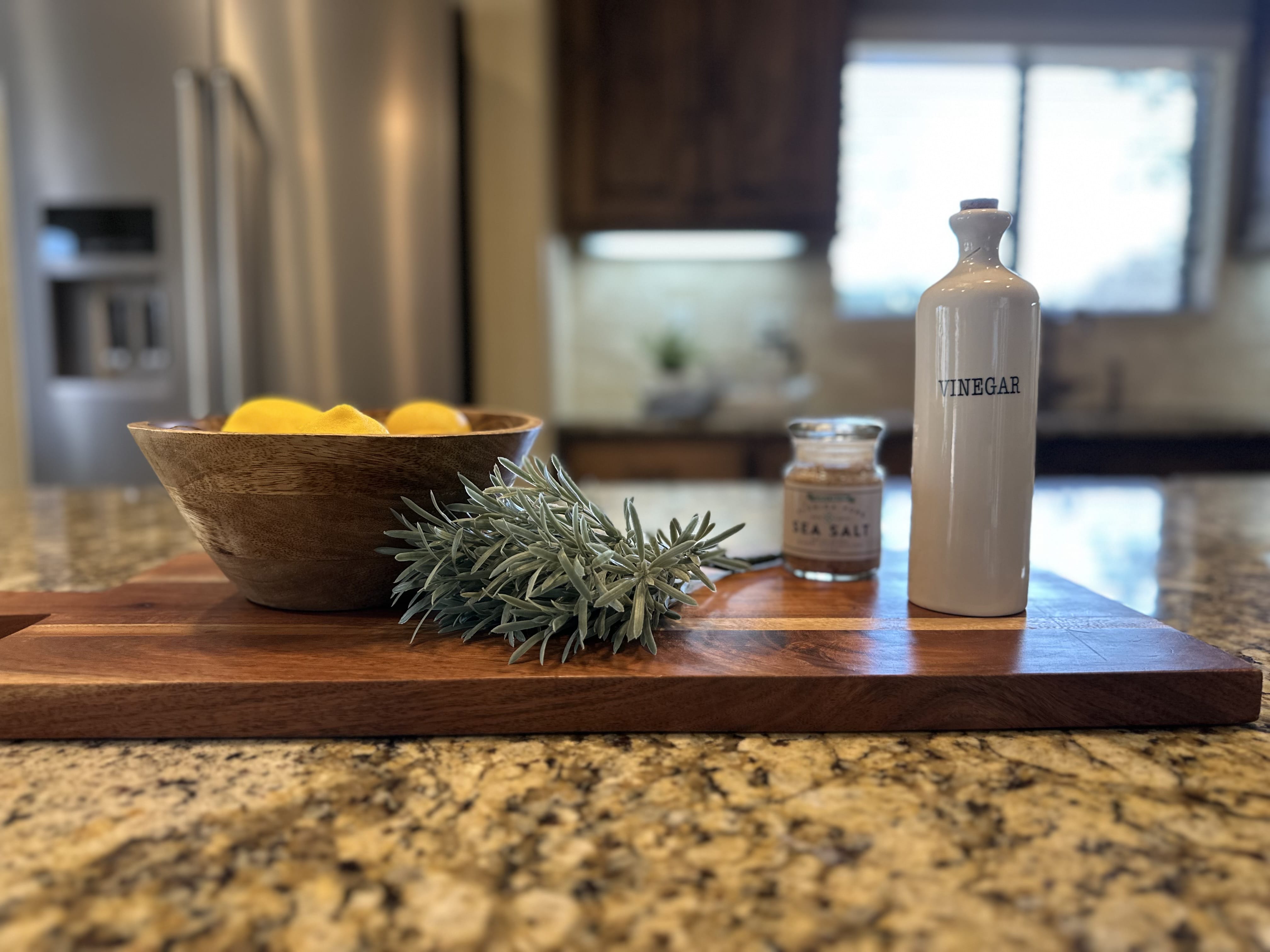 A close-up of a kitchen island countertop featuring a wooden cutting board styled with a bowl of lemons, a sprig of fresh rosemary, a small jar of sea salt, and a white ceramic vinegar bottle.