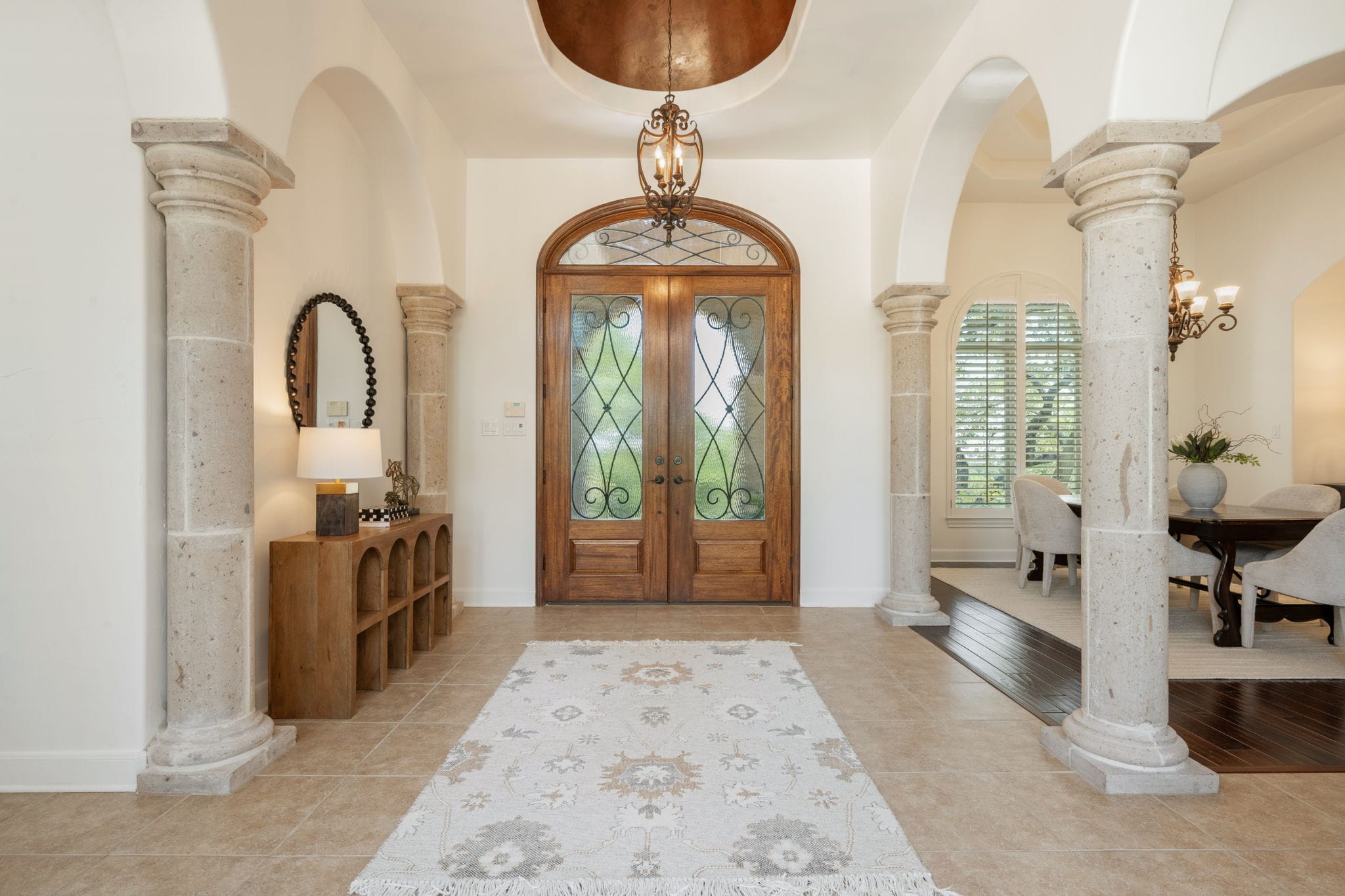 The grand interior entryway of a home features a high ceiling with a copper dome cutout and a hanging wrought-iron chandelier. Large, arched doorways are supported by prominent stone pillars with classic carved details. Double wooden front doors with ornate glass panels stand at the center. The floor is tiled with light-colored stone squares, accented by a patterned runner rug, leading into adjacent living spaces.