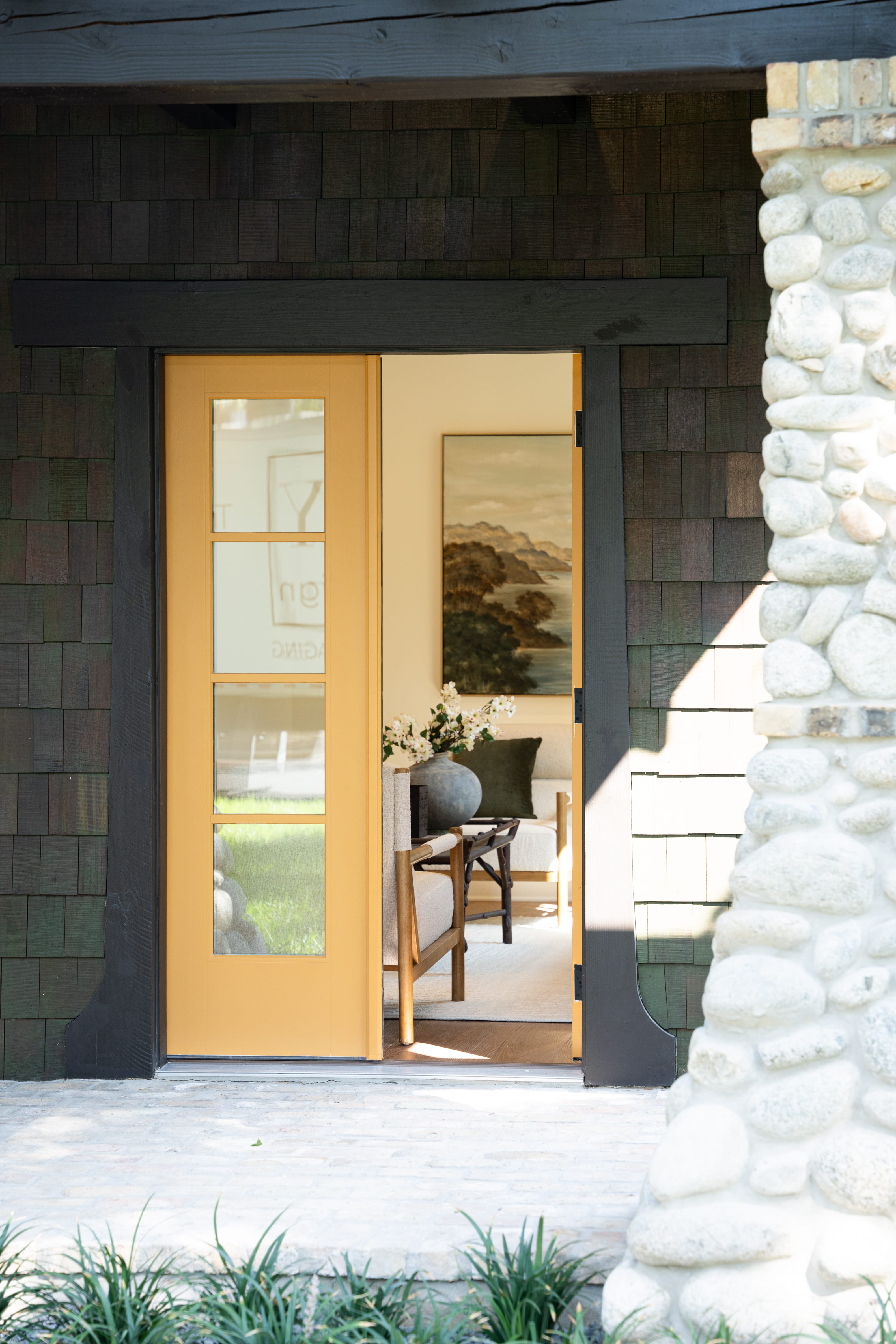 An exterior view looking through an open entryway into a cozy home. The doorway features a warm, mustard-yellow door with four glass panes, framed by thick black trim against dark wood shingle siding. To the right, a textured pillar made of light-colored river stones frames the porch. Through the open door, a glimpse of the interior reveals a well-lit living space with a wooden armchair, a large grey ceramic vase with white flowers on a side table, and a framed landscape painting of mountains and water hanging on the far wall.