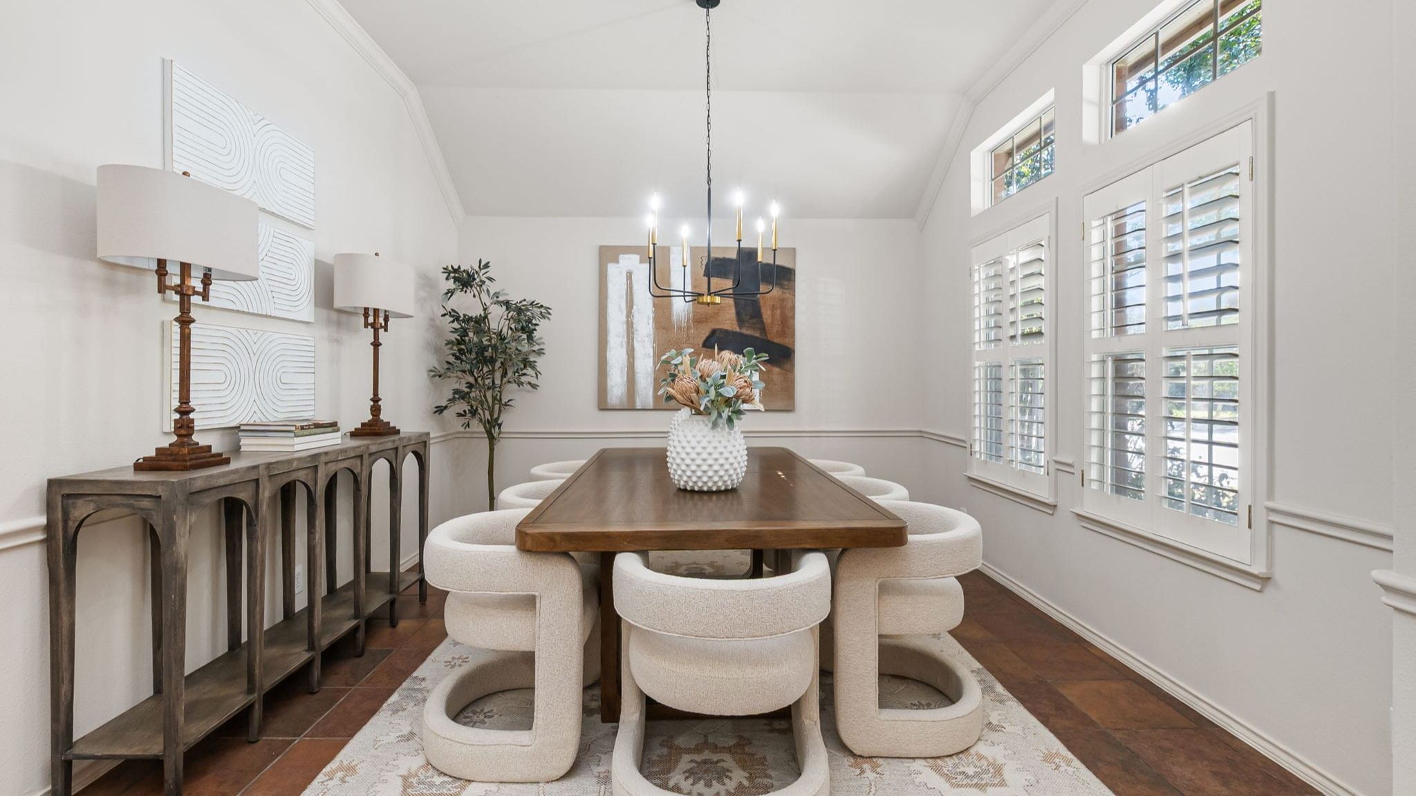 A modern dining room featuring a wooden table with white upholstered circular chairs. On the left, there is a wooden console table with two lamps. The walls are adorned with white textured artwork, and a large painting hangs above the table. The room is illuminated by a chandelier, and natural light filters through large windows with white shutters. The floor is covered with a patterned rug, and the overall color scheme is neutral, with earthy tones and soft lighting.