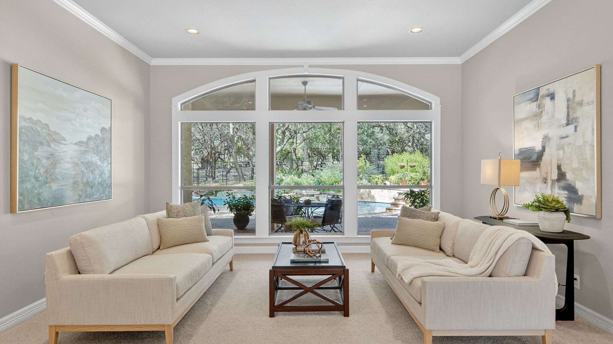Sunlit living room with two cream sofas facing each other, a wooden coffee table between them, large arched windows overlooking a patio and greenery, neutral walls, and modern accent lighting