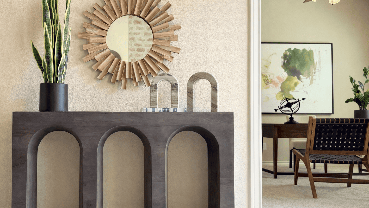 Stylish interior scene with a dark console table featuring arched details, topped with a round sunburst mirror, plants, and decor; beside it, a bright dining area with a wooden table, woven chairs, framed wall art, and greenery.