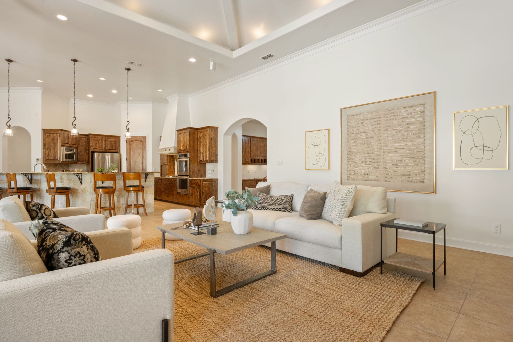 An open-concept living space highlights a bright sitting area seamlessly connecting to a kitchen in the background. In the living space, a long white sofa and matching armchairs flank a gray rectangular coffee table set on a large woven jute rug. The kitchen features dark wood cabinetry, stainless steel appliances, and a breakfast bar island lined with wooden barstools beneath hanging pendant lights. Framed abstract art pieces decorate the crisp white walls under a high, recessed tray ceiling.