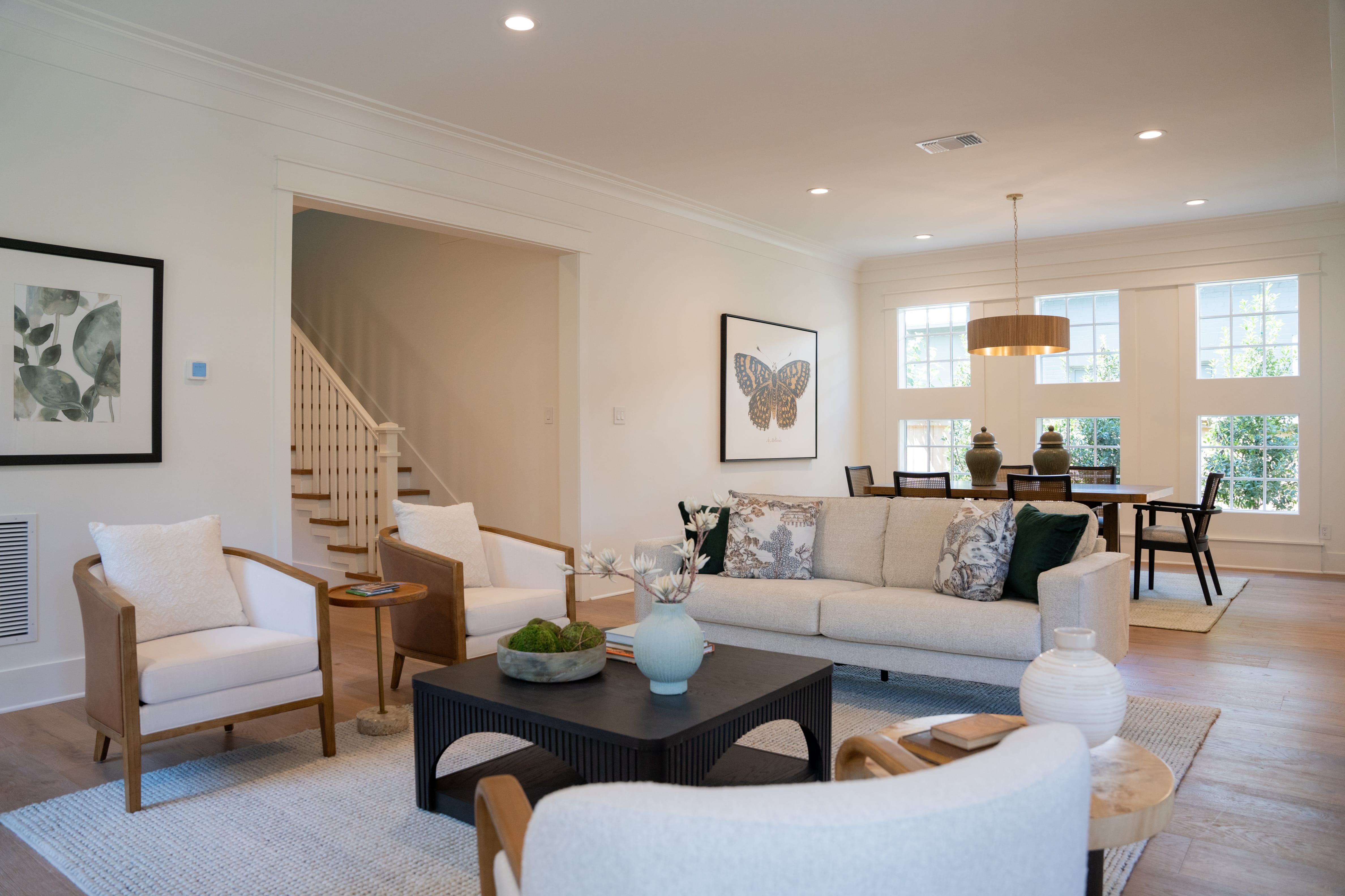 A spacious, open-concept living and dining area with bright white walls and light hardwood flooring. In the foreground, a modern living room setup features a cream-colored sofa styled with patterned and deep green pillows, facing a dark wood coffee table with a light blue vase and a bowl of green moss. Two leather-backed white armchairs sit to the left, alongside an arched doorway revealing a white staircase with wooden treads. On the back wall, a large framed sketch of a butterfly hangs above the sofa. In the background, a dining table with woven chairs sits under a drum-shaped wooden pendant light, positioned next to large grid windows that let in abundant natural light.