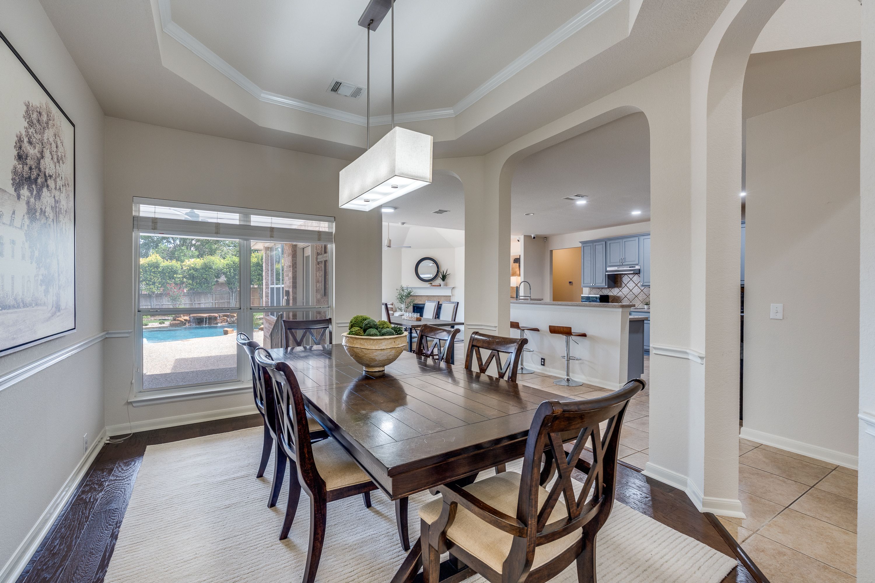 A spacious dining room featuring a wooden dining table with eight chairs, a decorative bowl in the center, and a modern light fixture hanging above. Large windows let in natural light, offering a view of the backyard. In the background, you can see a glimpse of the kitchen with light-colored cabinetry and an open floor plan