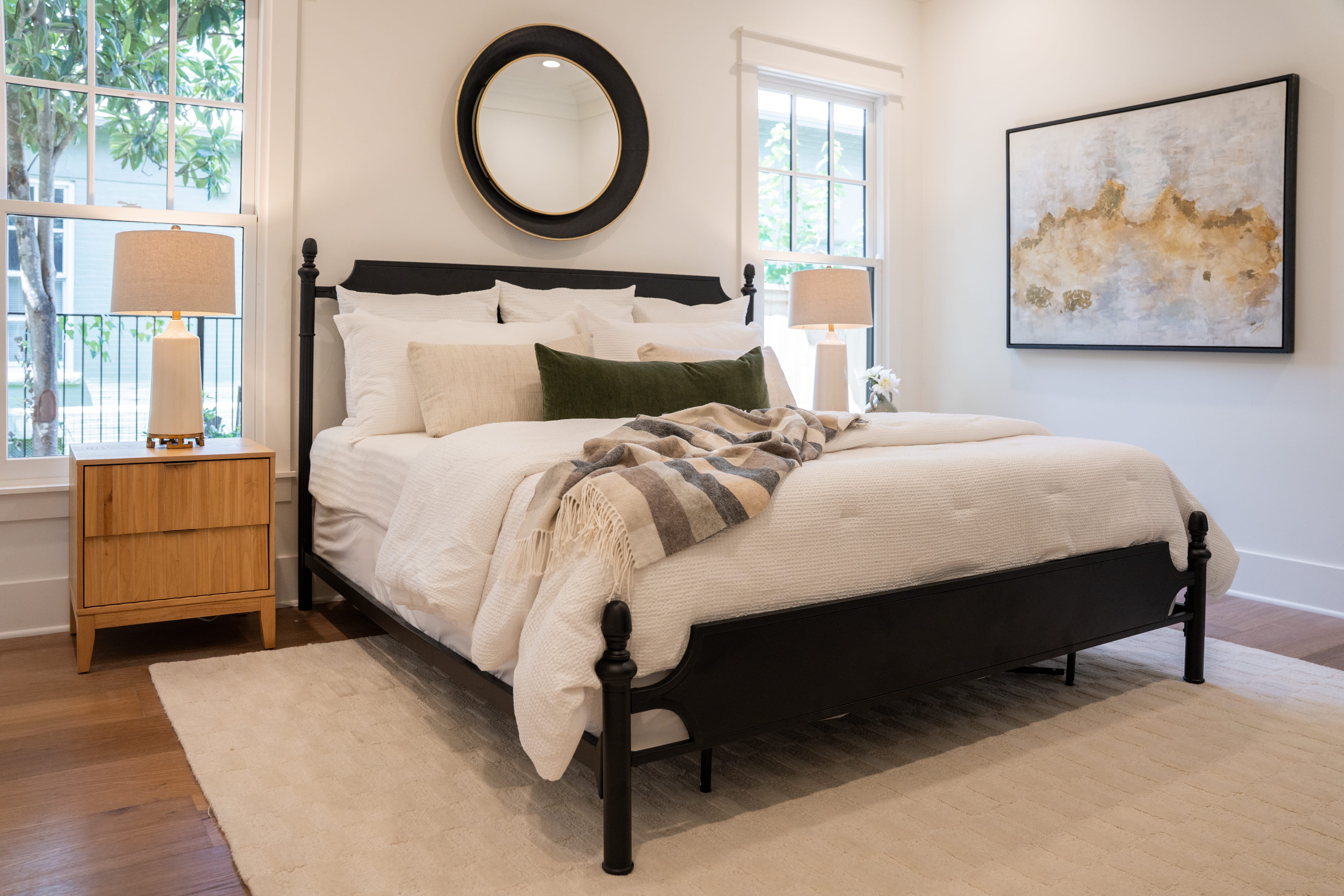 A bright and inviting bedroom featuring a black bed frame centered on a textured beige area rug over warm hardwood floors. The bed is styled with plush white bedding, a beige accent pillow, a long dark green velvet lumbar pillow, and a casual grey, tan, and white striped throw blanket draped near the foot. Above the headboard hangs a round mirror with a thick black and gold frame. To the left of the bed, a light wood two-drawer nightstand holds a white ceramic table lamp with a natural fabric shade. On the right wall, a large framed abstract painting in white, beige, and gold tones complements the room's serene, earthy color palette. Natural light streams in through two large grid windows.