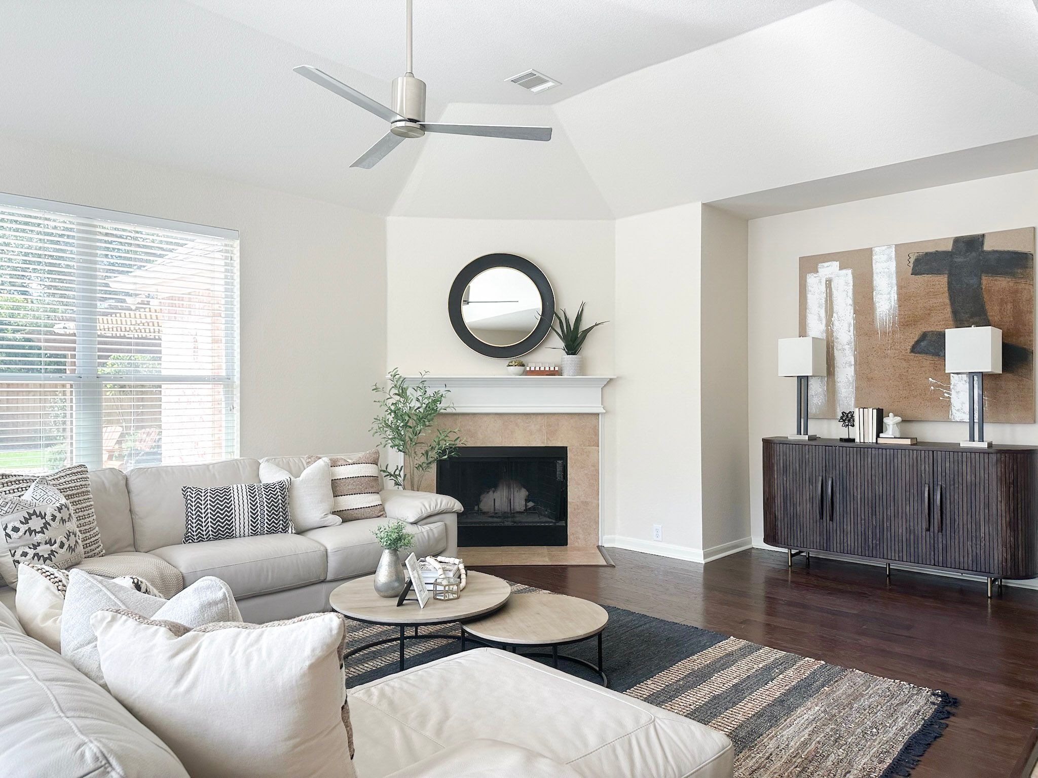 A cozy, well-decorated living room featuring a light beige sectional sofa, a round coffee table with minimalist decor, and a neutral-colored area rug. A fireplace is located in the background with a large mirror above it. Modern artwork hangs on the wall to the right, and natural light streams in through the windows on the left side of the room