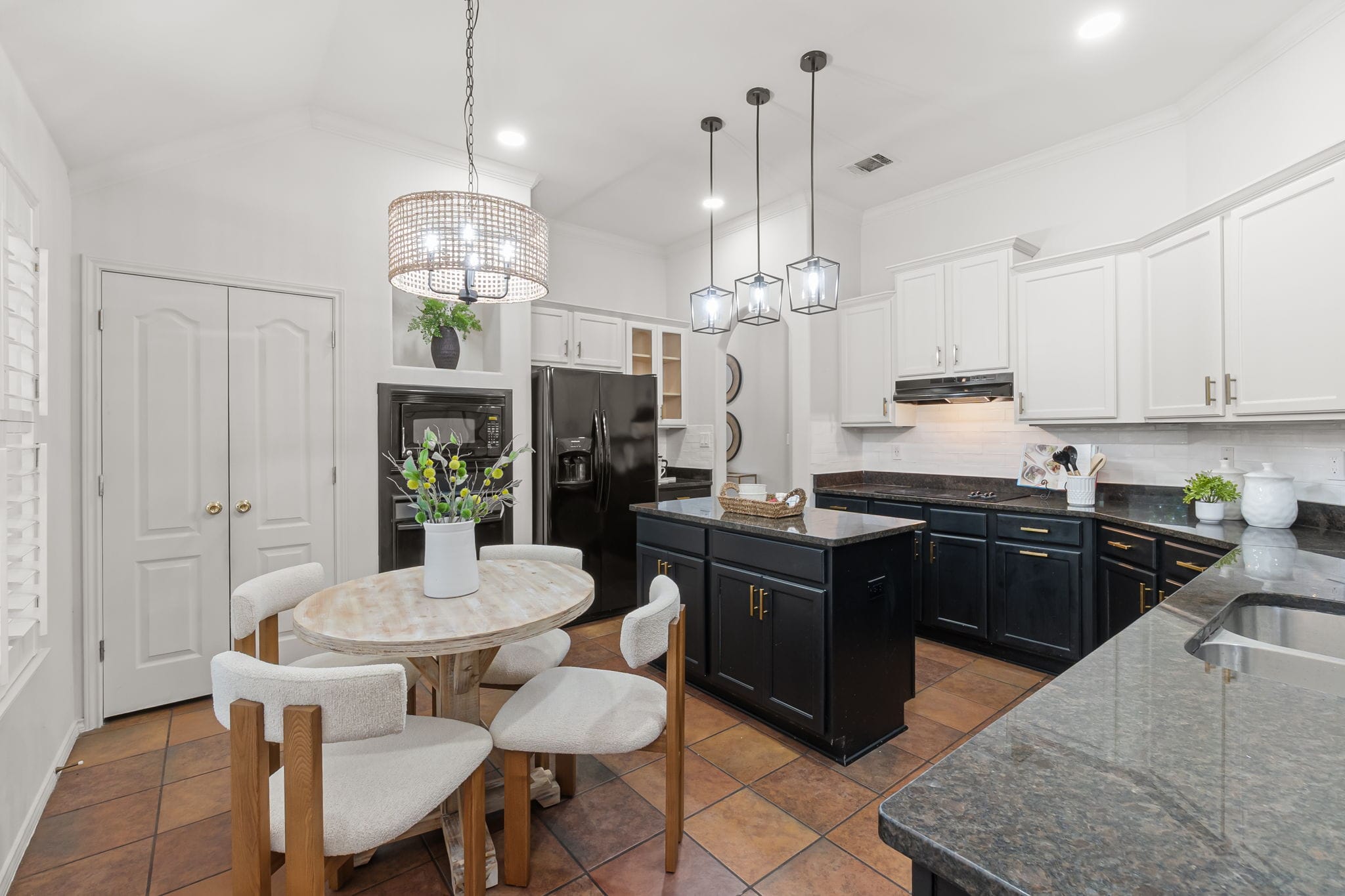 A bright and modern kitchen featuring a central wooden table with light-colored upholstered chairs. Above the table hangs a stylish woven pendant light. The kitchen has dark blue cabinets with gold handles, contrasting with the light countertops and backsplash. The black refrigerator and oven are located on the left side, while the kitchen island, with dark countertops, sits in the center. There are several plants and decorative items placed around the kitchen, adding a natural touch to the space. The floors are tiled with warm, earthy tones, and natural light enters through the window with white shutters.
