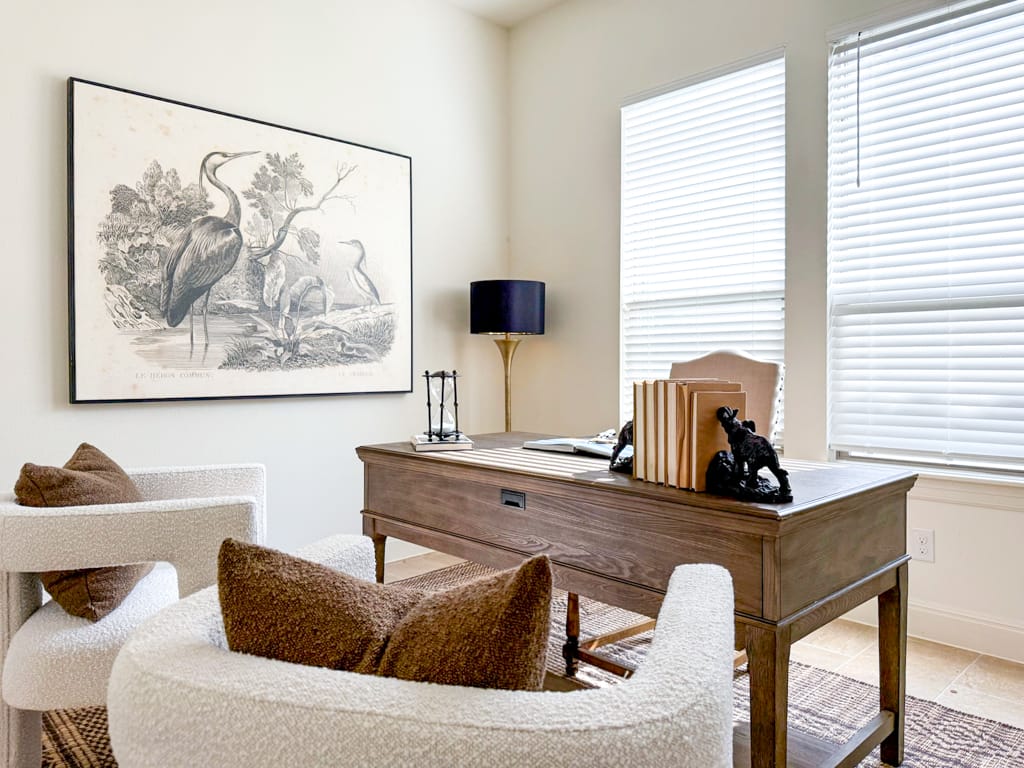Modern home office with a wooden desk, textured accent chairs, black table lamp, large bird artwork, and tall windows with white blinds creating a bright, airy workspace.