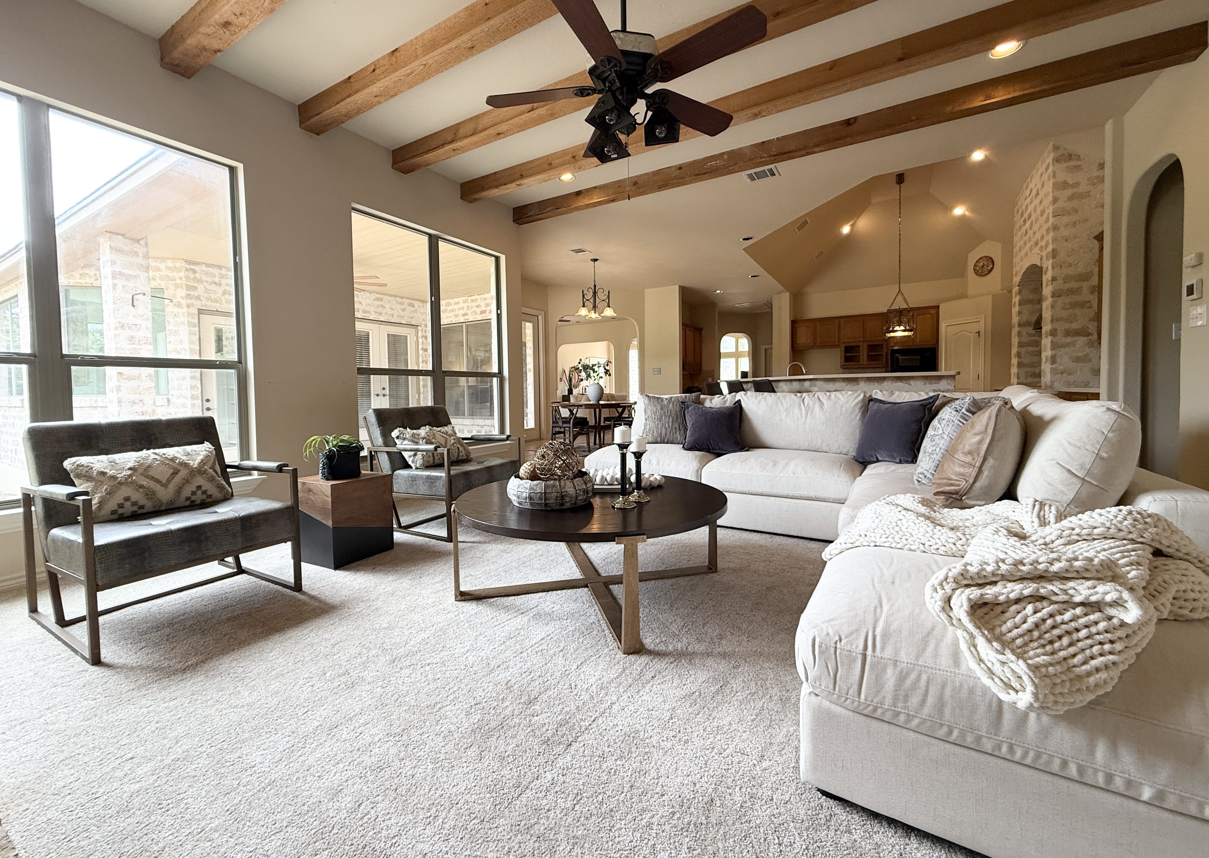 Helotes home staged living room featuring a large beige sectional, modern armchairs, round coffee table with decor, exposed wood beams, and open kitchen layout.