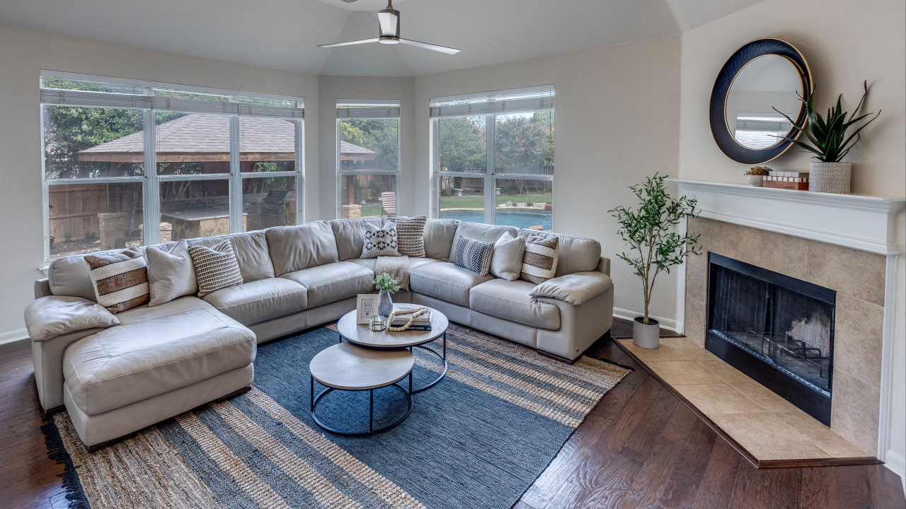 A spacious and well-lit living room featuring a large light-colored sectional sofa arranged in a corner with several throw pillows. The room has large windows offering views of the backyard and a pool. A round coffee table sits on a striped rug, and there is a fireplace with a plant and decorative items on the mantel. The ceiling features a modern fan, and the hardwood floors are a rich brown color.