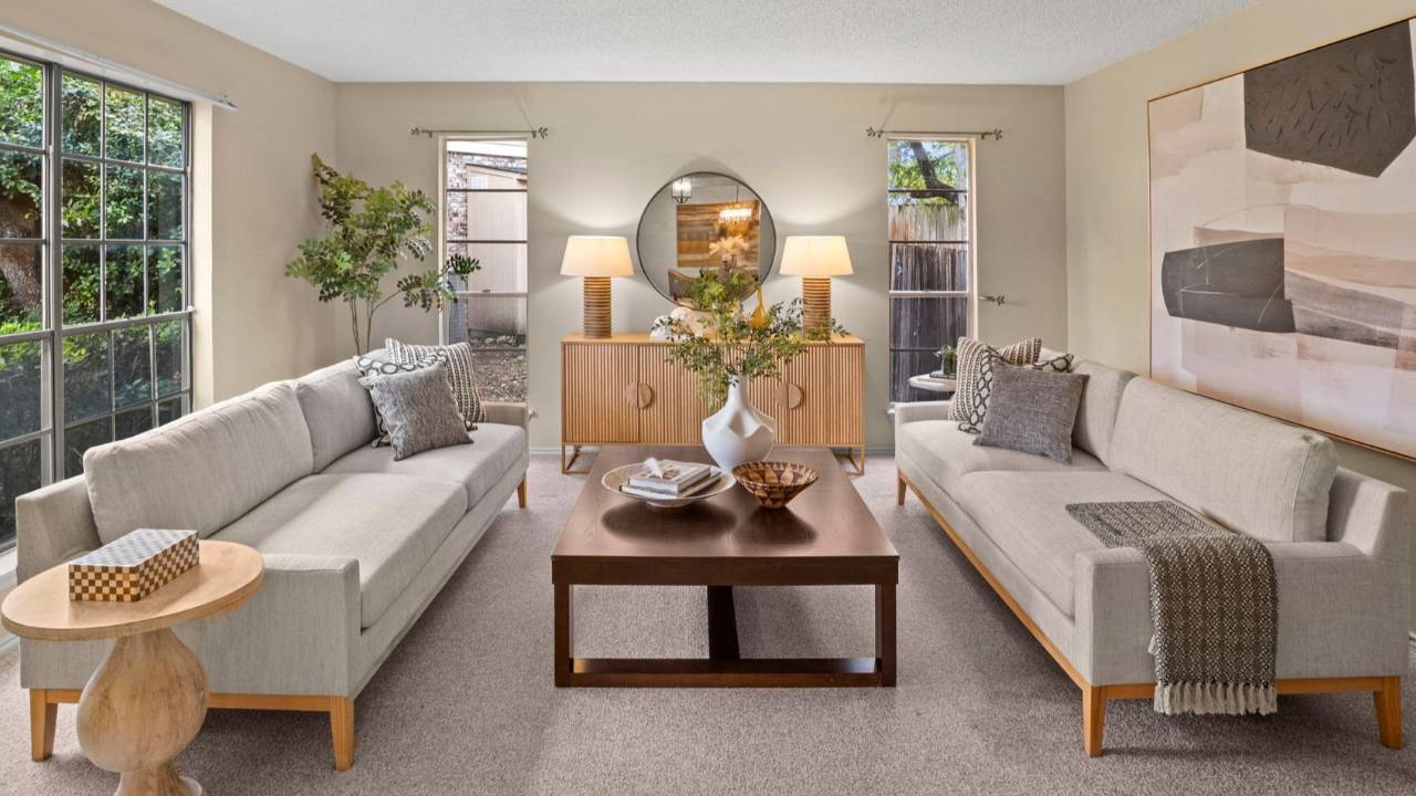 Bright, professionally staged living room with matching neutral sofas, wood coffee table, textured pillows and throws, modern sideboard with lamps, and large windows overlooking greenery.